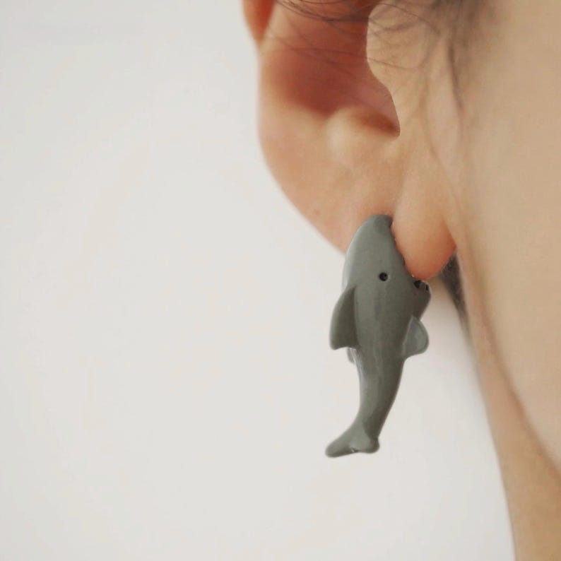 May include: A gray shark-shaped earring worn on an earlobe. The shark is depicted in a vertical orientation with its head pointing upward and its tail pointing downward, featuring a small black eye and visible dorsal and pectoral fins.