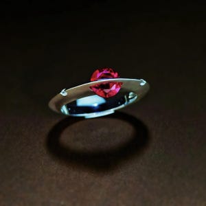 May include: A contemporary silver ring with a striking red gemstone. The gemstone is set in an open band design. The ring is presented against a dark backdrop, accentuating its elegant simplicity. A piece of jewellery.