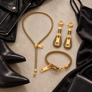 May include: A jewelry set consisting of a necklace, a bracelet, and a pair of drop earrings. Each piece is crafted from black zipper teeth with gold-toned metal accents, featuring a prominent gold zipper pull tab as a central design element.