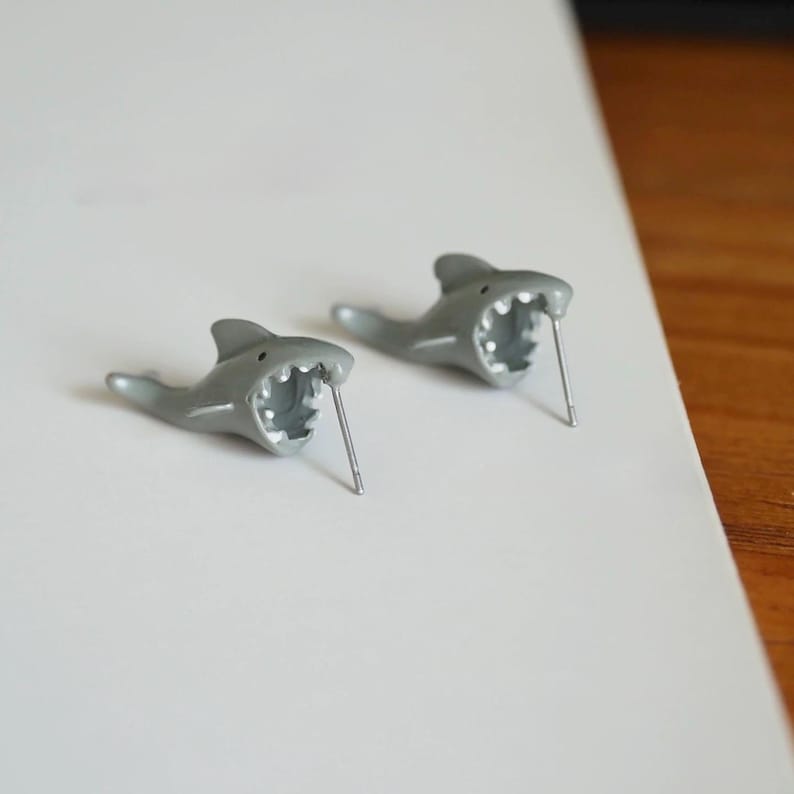 May include: A pair of gray shark-shaped stud earrings. Each earring features an open mouth with visible teeth and a silver post. The earrings are made of a smooth, matte material and are set against a white background.