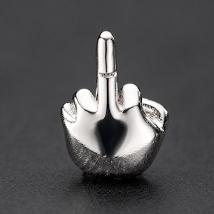 May include: A small, polished silver-toned metal charm shaped like a hand making a middle finger gesture. The object features a smooth, reflective surface with defined contours representing the fingers and knuckles in a compact, three-dimensional form.