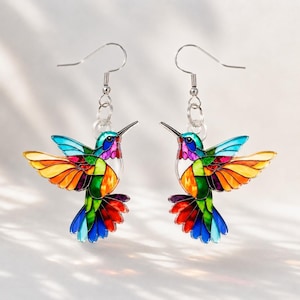 Peut inclure: Une paire de boucles d'oreilles pendantes en forme de colibri avec un effet vitrail. Les oiseaux sont composés de segments multicolores vifs en rouge, orange, jaune, vert, bleu et violet, suspendus à des crochets en métal argenté.