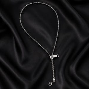 May include: A silver-toned necklace designed to resemble a zipper. The piece features a continuous chain of interlocking metal teeth forming a circular loop, connected to a rectangular zipper pull tab and a small lobster claw clasp.