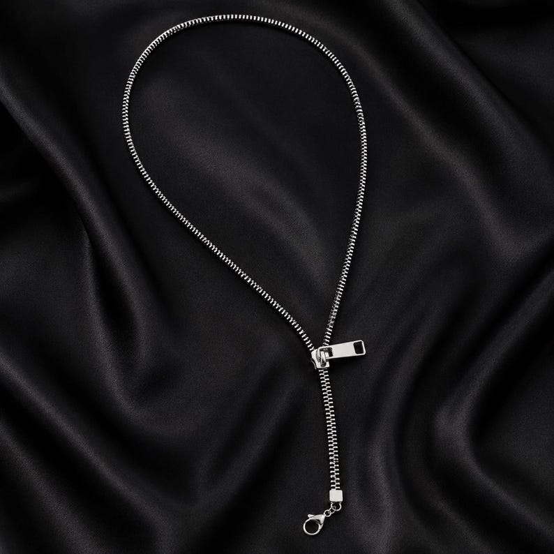 May include: A silver-toned necklace designed to resemble a zipper. The piece features a continuous chain of interlocking metal teeth forming a circular loop, connected to a rectangular zipper pull tab and a small lobster claw clasp.