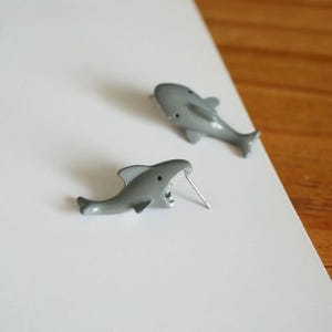May include: Two gray shark-shaped stud earrings. The earrings are detailed with open mouths and small black eyes. The earrings are on a white surface.