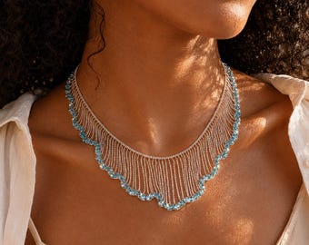 May include: A silver-toned fringe necklace featuring multiple delicate vertical chains that drape across the collarbone. The lower edge of the necklace is accented with a scalloped border of small, round, light blue gemstones set in metal.