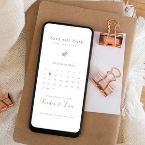 May include: A smartphone screen displaying a save the date announcement for a wedding in December 2023. The announcement features a calendar with the date highlighted, the couple's names "Kerstin & Liam", and the text "Save the Date, Wir Sagen Ja!" in German.