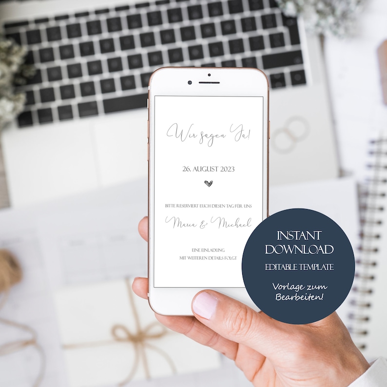 May include: A smartphone mockup displaying a wedding save the date announcement. The announcement is white with black text and a small heart icon. The text reads "Wir sagen Ja!" (We say Yes!), "26. August 2023", "Bitte reserviert euch diesen Tag f&uuml;r uns", "Maria & Michael", "Eine Einladung mit weiteren Details folgt". The background is a blurred image of a laptop keyboard and a notebook.