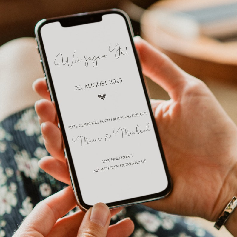 May include: A smartphone screen displaying a wedding announcement. The text reads "Wir sagen Ja!" (We say Yes!), "26. AUGUST 2023", "BITTE RESERVIERT EICH DIESEN TAG F&Uuml;R UNS" (Please reserve this day for us), "Maria & Michael", "EINE EINLADUNG" (An invitation), and "MIT WEITEREN DETAILS FOLGT" (With further details to follow).