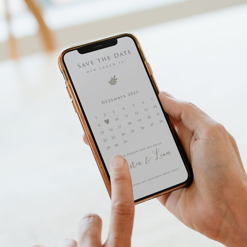 May include: A smartphone displaying a save the date announcement for a wedding in December 2023. The announcement features a calendar with a heart icon marking the date, the text "Save the Date", "Wir sagen Ja!" and "Justin & Lian".