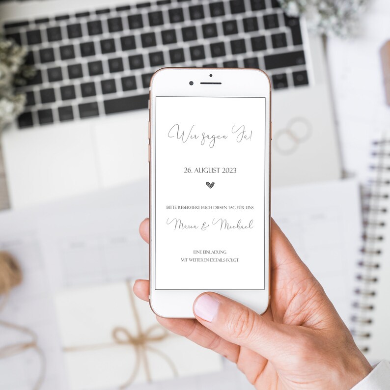 May include: A smartphone displaying a wedding announcement with the text "Wir sagen Ja!" (We say Yes!), August 26, 2023, and the names "Mana & Michael".
