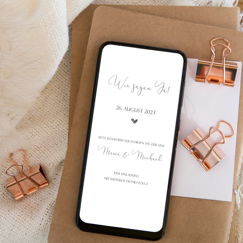 May include: A smartphone screen displaying a wedding save the date announcement. The text reads "Wir sagen Ja!" (We say Yes!), "26. AUGUST 2023", "Bitte reserviert euch diesen Tag f&uuml;r uns" (Please reserve this day for us), "Maria & Michael", "Eine Einladung mit weiteren Details folgt" (An invitation with further details will follow).