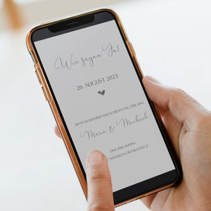 May include: A smartphone screen displaying a wedding announcement. The text reads "Wir sagen Ja!" (We say Yes!), "26. AUGUST 2023", "BITTE RESERVIERT EUCH DIESEN TAG F&Uuml;R UNS" (Please reserve this day for us), "Maria & Michael", and "EINE EINLADUNG MIT WEITEREN DETAILS FOLGT" (An invitation with further details will follow).