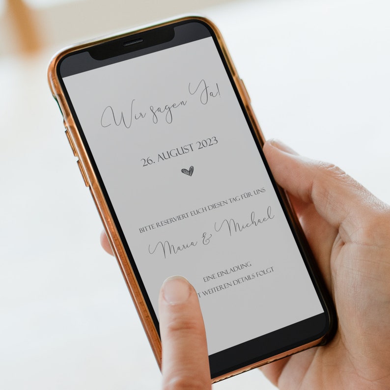 May include: A smartphone screen displaying a wedding announcement. The text reads "Wir sagen Ja!" (We say Yes!), "26. AUGUST 2023", "BITTE RESERVIERT EUCH DIESEN TAG F&Uuml;R UNS" (Please reserve this day for us), "Maria & Michael", and "EINE EINLADUNG MIT WEITEREN DETAILS FOLGT" (An invitation with further details will follow).