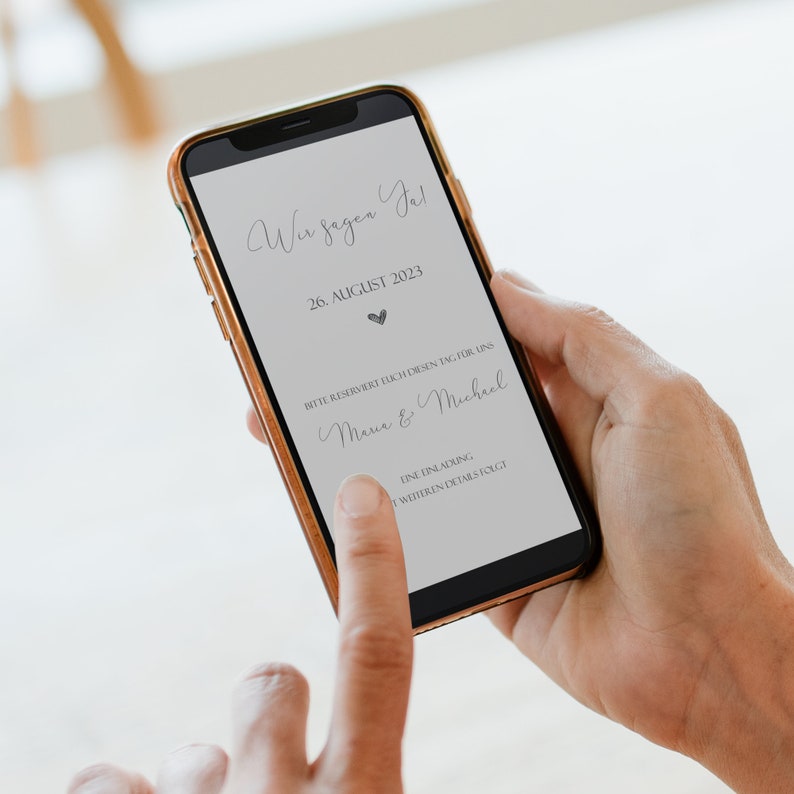 May include: A smartphone screen displaying a wedding announcement. The announcement reads "Wir sagen Ja!" (We say yes!), August 26, 2023, and includes the names "Maria & Michael".