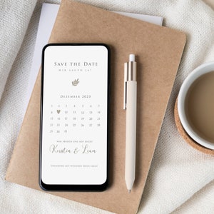 May include: A smartphone screen displaying a save the date announcement for a wedding in December 2023. The announcement features a calendar with a heart marking the date, the text "Save the Date", "Wir sagen Ja!" and "Wir freuen uns auf euch!" and the names "Kerstin & Liam".