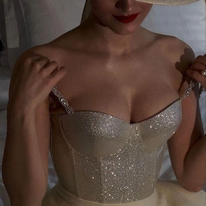 May include: A woman wearing a wide-brimmed, cream-coloured hat with a satin ribbon and a sparkling, silver bustier. The bustier has delicate, jewelled straps. The woman has red lipstick and is holding the straps of the bustier.