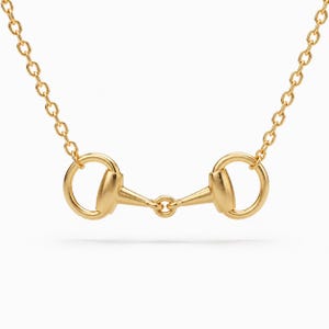 May include: A gold-coloured necklace featuring a central horse bit pendant. The pendant consists of two circular rings connected by a horizontal bar with a small central ring. The piece is suspended from a delicate gold-coloured chain with oval links.