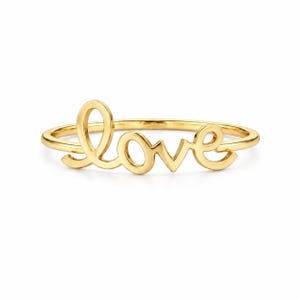 May include: A gold ring spelling out the word "love" in a cursive script. The ring features a delicate band with the letters of "love" joined together. The ring is set against a plain white backdrop.