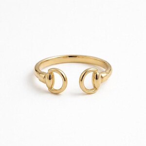 Pode incluir: Um anel dourado com um design aberto. O anel apresenta um design de freio de cavalo na frente, com dois círculos abertos e arredondados conectados à banda. O anel está sobre um fundo branco.