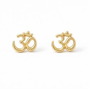 May include: A pair of gold stud earrings, each featuring a polished, three-dimensional Om symbol. The earrings have a smooth, reflective metallic finish and a rounded, sculptural design that captures light across the curved surfaces of the emblem.