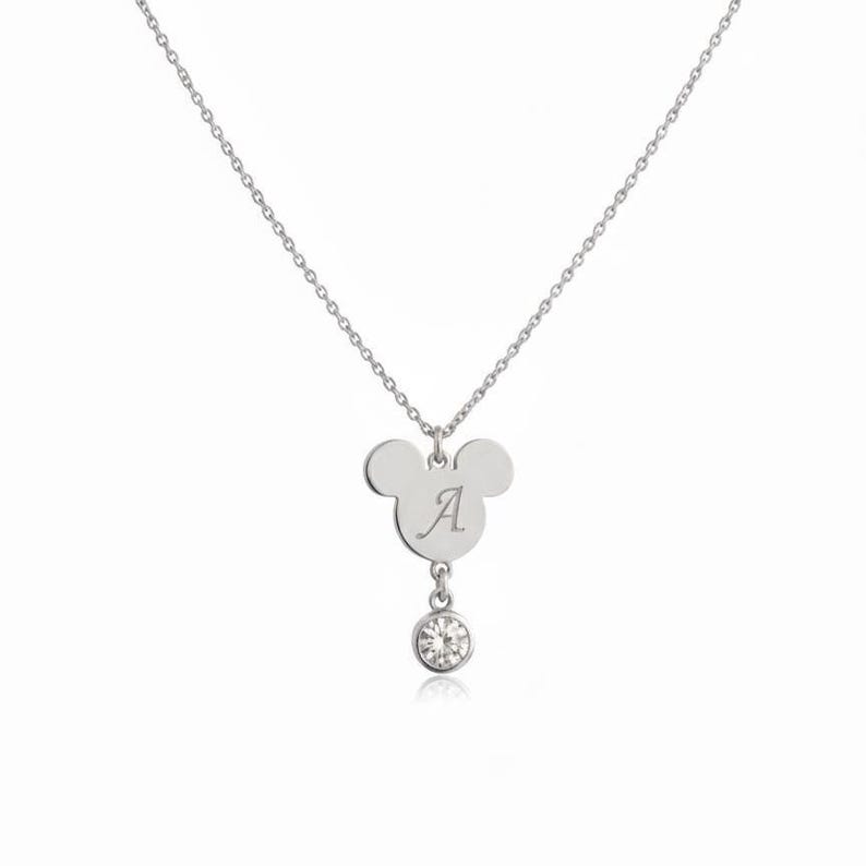 Peut inclure: Un collier argent&eacute; avec un pendentif en forme d'oreilles de Mickey Mouse, grav&eacute; de la lettre "A". Une petite pierre pr&eacute;cieuse ronde et transparente est suspendue en dessous. La cha&icirc;ne du collier est &eacute;galement argent&eacute;e.