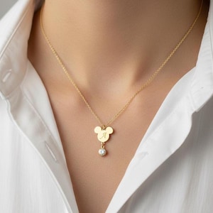 Peut inclure: Collier dor&eacute; avec un pendentif en forme de Mickey Mouse. Le pendentif porte la lettre "A" grav&eacute;e et un petit bijou rond et transparent en dessous. Le collier est port&eacute; sur une chemise blanche.