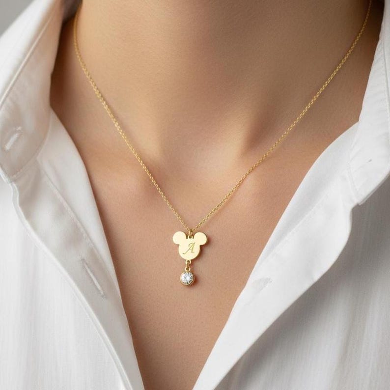 Peut inclure: Collier dor&eacute; avec un pendentif en forme de Mickey Mouse. Le pendentif porte la lettre "A" grav&eacute;e et un petit bijou rond et transparent en dessous. Le collier est port&eacute; sur une chemise blanche.