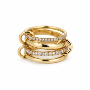 May include: Four gold rings interlocked with small loops. Two of the rings are encrusted with small, clear gemstones. The rings are a bright, shiny gold colour and are set against a white background.