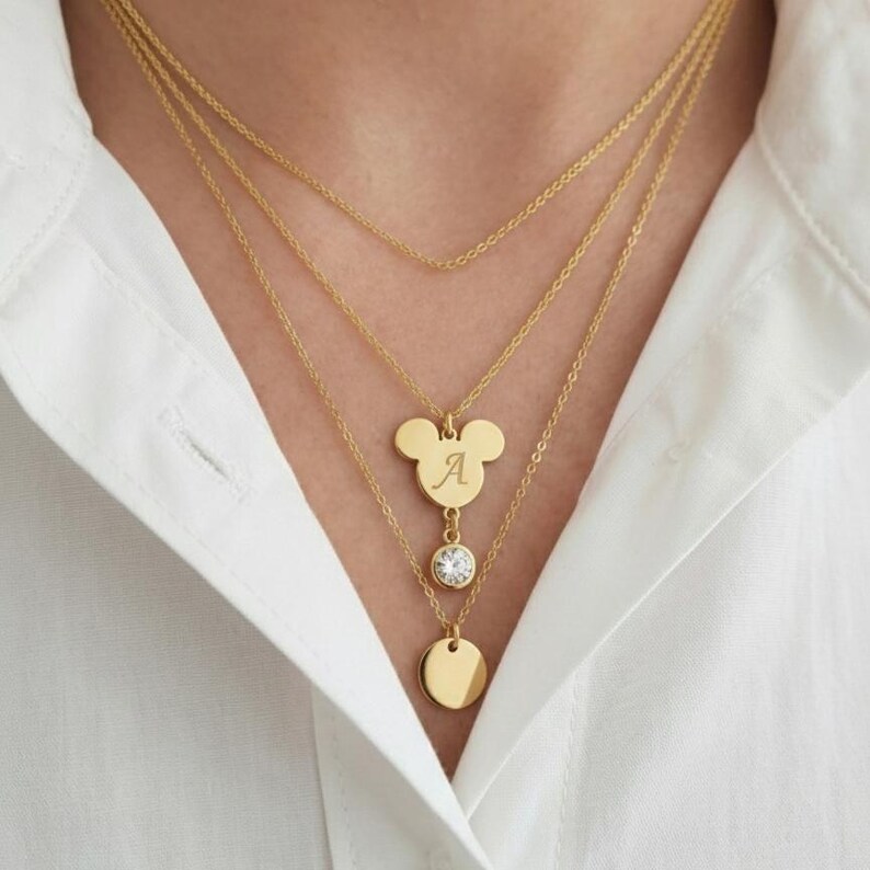 Peut inclure: Un ensemble de colliers dor&eacute;s superpos&eacute;s. La couche sup&eacute;rieure est une cha&icirc;ne d&eacute;licate. La deuxi&egrave;me couche a un pendentif en forme de Mickey Mouse avec la lettre "A". La troisi&egrave;me couche a un pendentif rond en cristal clair. La couche inf&eacute;rieure a un petit pendentif rond.