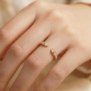 May include: A gold open-style ring featuring two pointed ends, each set with three small, clear, round-cut stones. The band is polished gold, and the design leaves a gap between the two decorative ends, creating a bypass or wrap-around appearance.