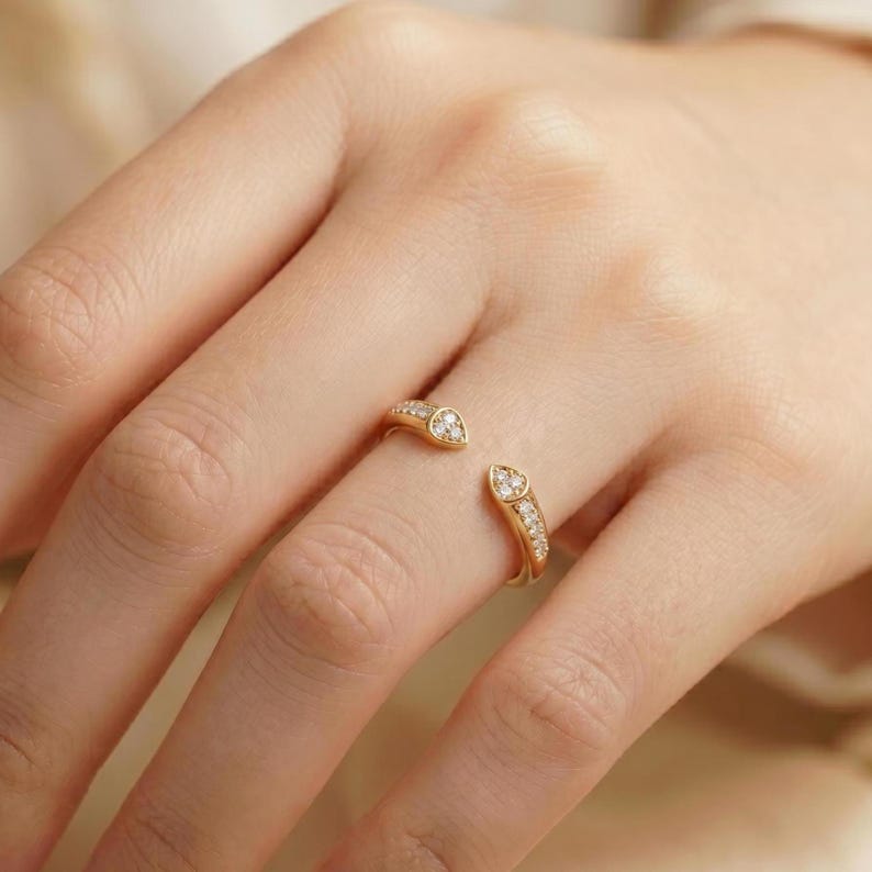 May include: A gold open-style ring featuring two pointed ends, each set with three small, clear, round-cut stones. The band is polished gold, and the design leaves a gap between the two decorative ends, creating a bypass or wrap-around appearance.