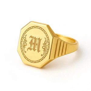 以下が含まれることがあります： 八角形の面を持つゴールドカラーのシグネットリング。表面には、装飾的な円形のボーダーの中に彫刻された文字「M」があります。リングは研磨仕上げで、バンドには溝のあるデザインが施されています。