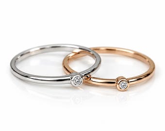 May include: Two thin metal rings, one in silver-toned metal and one in rose gold-toned metal, each featuring a single small, round, clear gemstone set in a bezel setting at the center of the band, creating a simple and elegant jewelry design.