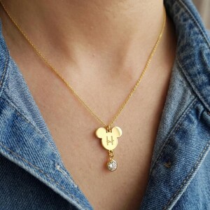 Peut inclure: Collier pendentif dor&eacute; avec un charm en forme de Mickey Mouse. Le charm porte la lettre "H" grav&eacute;e et un cristal clair pendant. Le collier est port&eacute; sur une veste en jean.