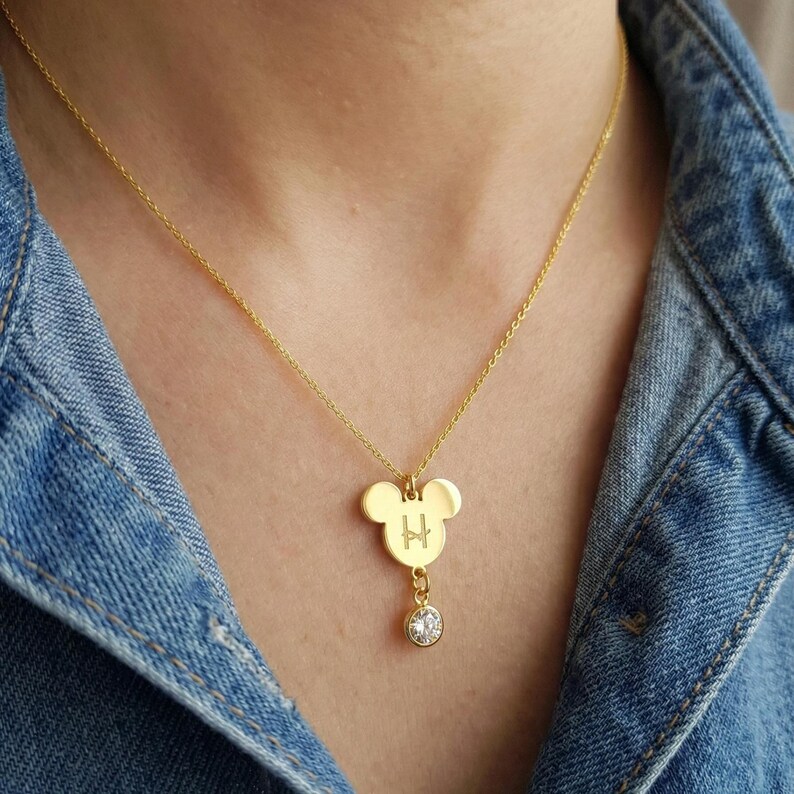 Peut inclure: Collier pendentif dor&eacute; avec un charm en forme de Mickey Mouse. Le charm porte la lettre "H" grav&eacute;e et un cristal clair pendant. Le collier est port&eacute; sur une veste en jean.