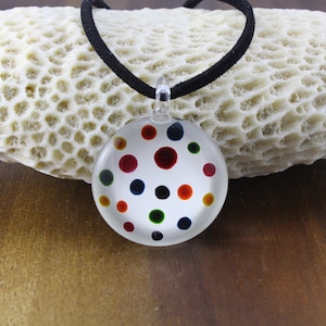 Peut inclure: Un collier avec un pendentif rond en verre sur un cordon noir. Le pendentif est transparent et présente des pois colorés en rouge, orange, jaune, vert, bleu et noir.