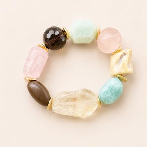 Könnte beinhalten: Ein elastisches Armband mit einer Vielzahl von verschiedenfarbigen, unregelmäßig geformten und facettierten Edelsteinperlen. Die Perlen in Hellrosa, Hellblau, Dunkelbraun, durchscheinendem Gelb und Creme sind durch kleine goldfarbene Metallzwischenstücke getrennt.