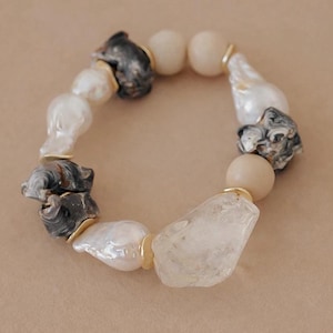 Könnte beinhalten: Ein Perlenarmband mit einer Mischung aus unregelmäßigen weißen Barockperlen, dunkelgrauen marmorierten Steinperlen, glatten beigen runden Perlen und einem einzelnen klaren, gezackten kristallartigen Stein, getrennt durch kleine goldfarbene Metallabstandshalter.