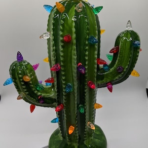 May include: A ceramic cactus Christmas decoration with a glossy green finish. The cactus is adorned with colorful, faceted lights and topped with a yellow star. A festive and unique holiday accent.