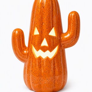 May include: A glossy orange cactus-shaped ceramic decoration with a speckled finish and a carved jack-o'-lantern face. The interior is illuminated, casting a warm light through the triangular eyes and the jagged, smiling mouth cutouts.