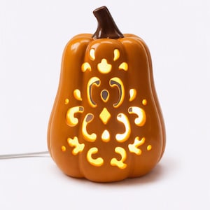 May include: A glossy orange ceramic pumpkin lamp with a dark brown stem. The pumpkin features intricate, symmetrical laser-cut patterns that allow warm, glowing light to shine through from the interior, connected by a thin white electrical cord.