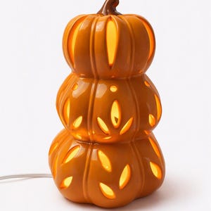 May include: A stack of three orange ceramic pumpkins forming a decorative lamp. The pumpkins feature cut-out leaf-shaped patterns that emit a warm, glowing light from within. A brown stem sits on top, and a white electrical cord extends from the base.