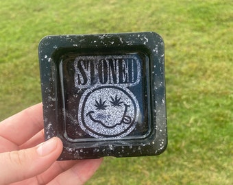 Op de afbeelding: Zwarte en witte harsbak met een glitterafwerking. De bak heeft een smiley-gezicht met het woord "STONED" erboven.