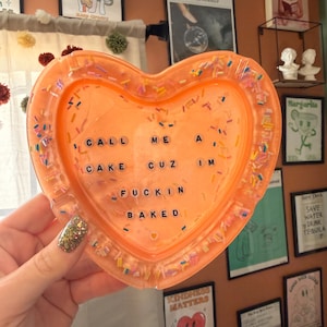 Puede incluir: Una bandeja de resina naranja en forma de corazón con chispas de colores y el texto "I CALL ME A CAKE CUZ I'M FUCKIN BAKED". La bandeja está sostenida por una mano con esmalte de uñas brillante. Hay objetos decorativos en el fondo.