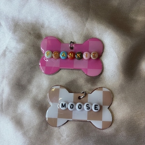 Puede incluir: Dos etiquetas para mascotas con forma de hueso. Una es rosa con un patrón de cuadros y el nombre "Frankie" escrito con cuentas de colores. La otra es de color beige y blanco a cuadros con el nombre "Moose" en cuentas blancas.