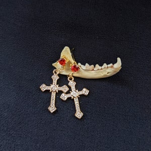 May include: A pair of gold cross earrings with red crystal accents. The earrings are dangling and have a delicate design.