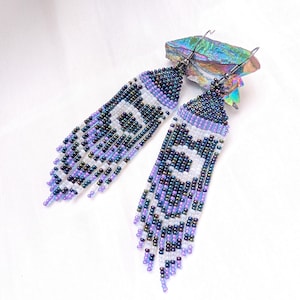 May include: A pair of long, dangling earrings crafted from small glass beads in shades of iridescent dark blue, purple, and white. The beads are arranged in a geometric pattern with a fringe at the bottom, attached to silver-toned metal hooks.