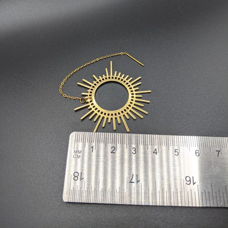 May include: Gold sunburst dangle earring with a chain and a small bead. The earring measures approximately 3 inches in diameter.
