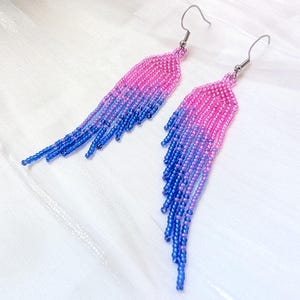 May include: A pair of long, dangling earrings crafted from small seed beads. The design features a vibrant color gradient, transitioning from bright pink at the top to a deep blue at the bottom, with individual beaded strands creating a fringe effect.